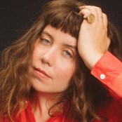 A woman wearing a red shirt with her hand rested on her head. She has a large gold coin ring on her pinky finger. She has a curly brown bob with a fringe